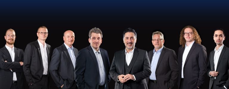 Gruppe von acht Männern in Anzügen vor schwarzem Hintergrund, in der Mitte E+H CEO Daniel Raseghi