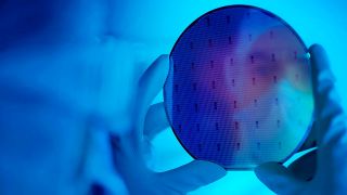 Hand mit Handschuh hält einen runden Siliziumwafer mit sichtbaren Strukturen vor blauem Hintergrund