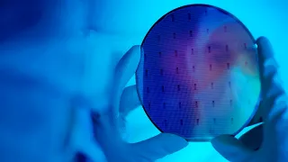 Hand mit Handschuh hält einen runden Siliziumwafer mit sichtbaren Strukturen vor blauem Hintergrund