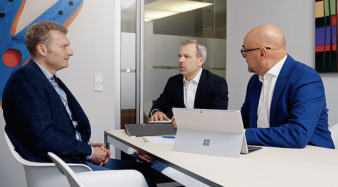 Die Drei Rechtsanwälte, Sommerfeld, Suntum und Frick in Anzügen sitzen an einem Konferenztisch in einem modernen Büro mit Glaswand und bunter Wandkunst