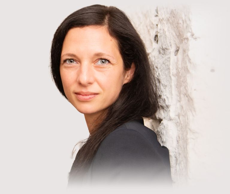 Sibylle Pöhler von Wein&Lese mit langen dunklen Haaren steht seitlich vor einer hellen Wand - DART