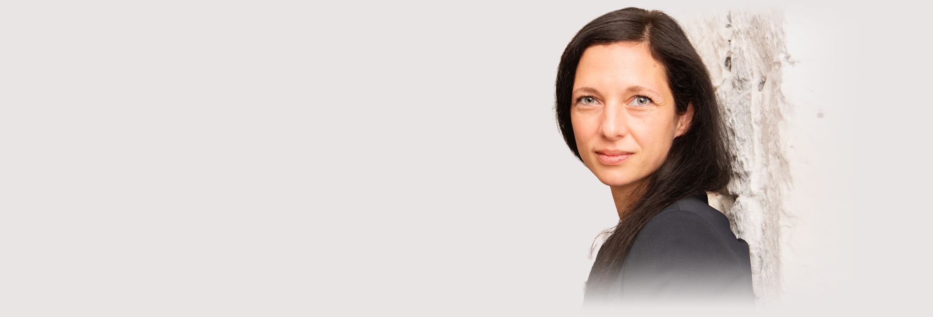 Sibylle Pöhler von Wein&Lese mit langen dunklen Haaren steht seitlich vor einer hellen Wand - DART